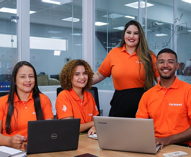 quatro pessoas em um ambiente corporativo, vestindo camisas laranjas com a marca da Brisanet. Três delas estão sentadas à mesa com notebooks abertos, enquanto uma mulher está em pé atrás delas, sorrindo.