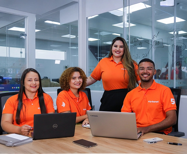 quatro pessoas em um ambiente corporativo, vestindo camisas laranjas com a marca da Brisanet. Três delas estão sentadas à mesa com notebooks abertos, enquanto uma mulher está em pé atrás delas, sorrindo.