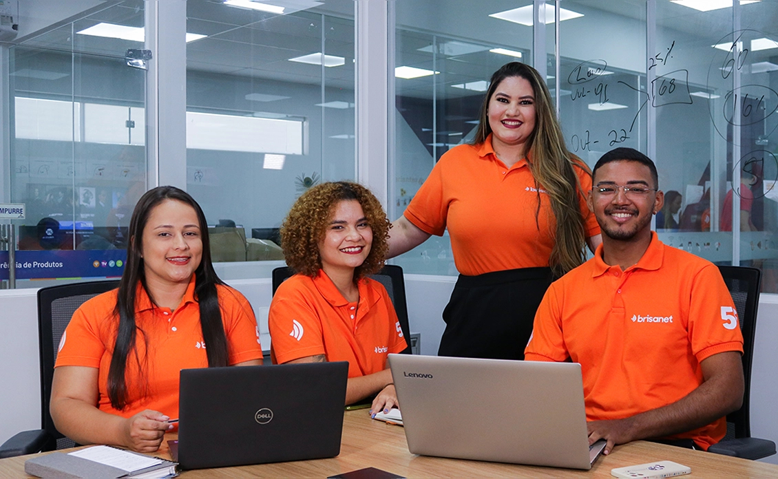 quatro pessoas em um ambiente corporativo, vestindo camisas laranjas com a marca da Brisanet. Três delas estão sentadas à mesa com notebooks abertos, enquanto uma mulher está em pé atrás delas, sorrindo.