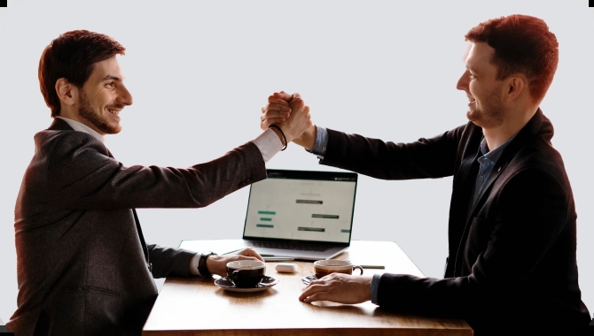 Dois homens em um ambiente de trabalho, vestindo ternos, sentados frente a frente em uma mesa com duas xícaras de café e um laptop ao fundo. Eles estão sorrindo e apertando as mãos. 