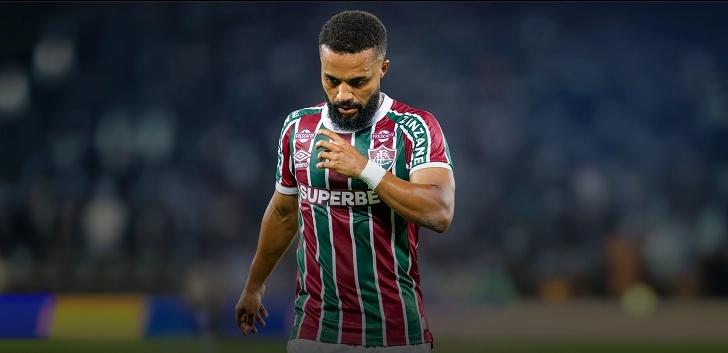 Jogador do Fluminense aparece em campo, veste uniforme tricolor do clube, ele esta olhando para baixo, o fundo da imagem é o estádio borrado.